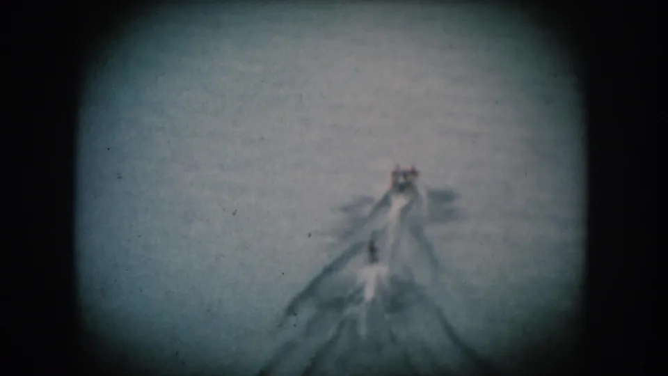 A single motorboat moves across open water leaving a pronounced V-wake; blue-shifted 8mm home movie footage consistent with mid-1950s American leisure.