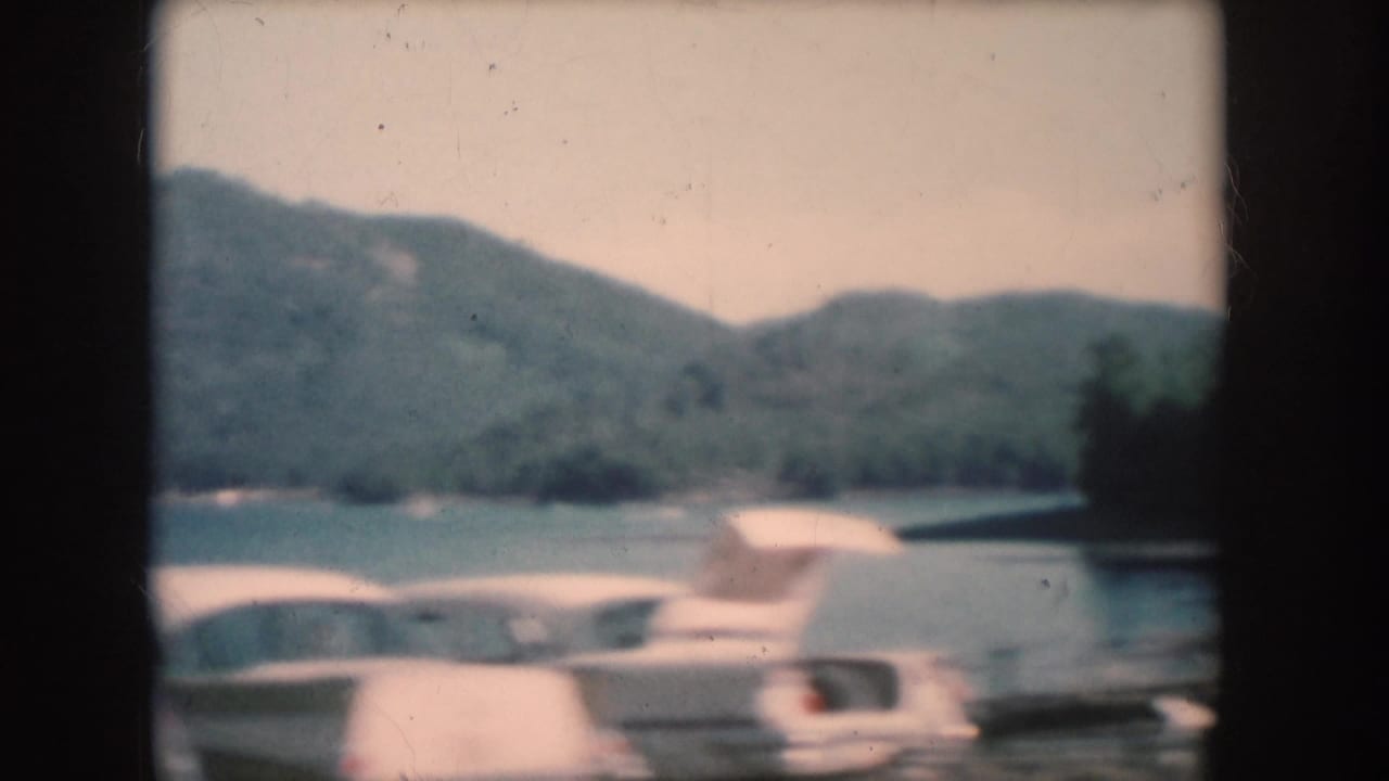 8mm color home movie frame of a lake with forested hills across the water and what appears to be a light-colored boat near shore, South Dakota, circa 1956.