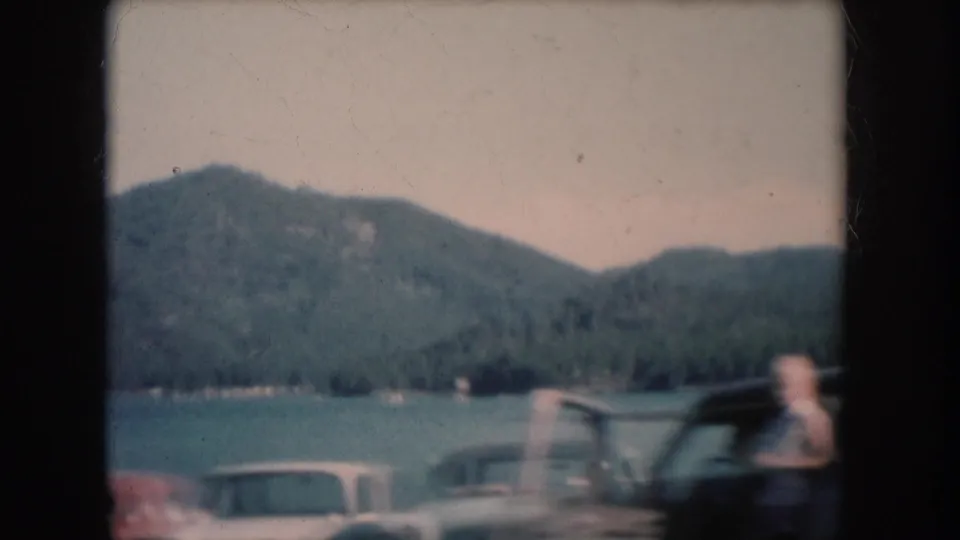 8mm home movie frame showing a lakeside parking area with dark-bodied vintage sedans and a blurred figure near vehicles; forested hills and lake visible in background, South Dakota, circa 1956.