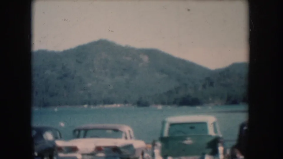 8mm color home movie of a lakeside parking area with several 1950s-era automobiles, including a white or cream-colored vehicle, forested hills and lake in background, South Dakota, circa 1956.