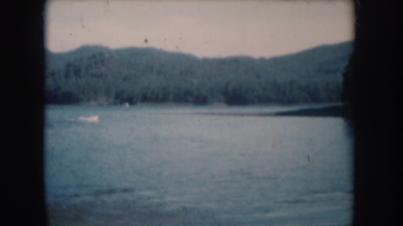 Calm lake with densely forested hills in background, filmed on 8mm with visible grain and faded color.