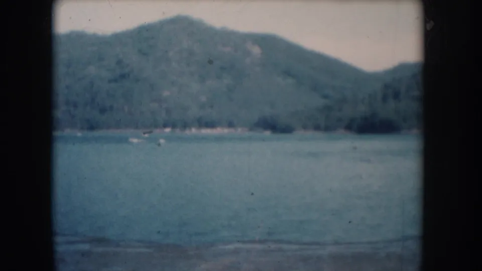 Forested hills above calm lake in the final frame of a 1956 8mm home movie attributed to South Dakota; small indistinct objects near far shore.