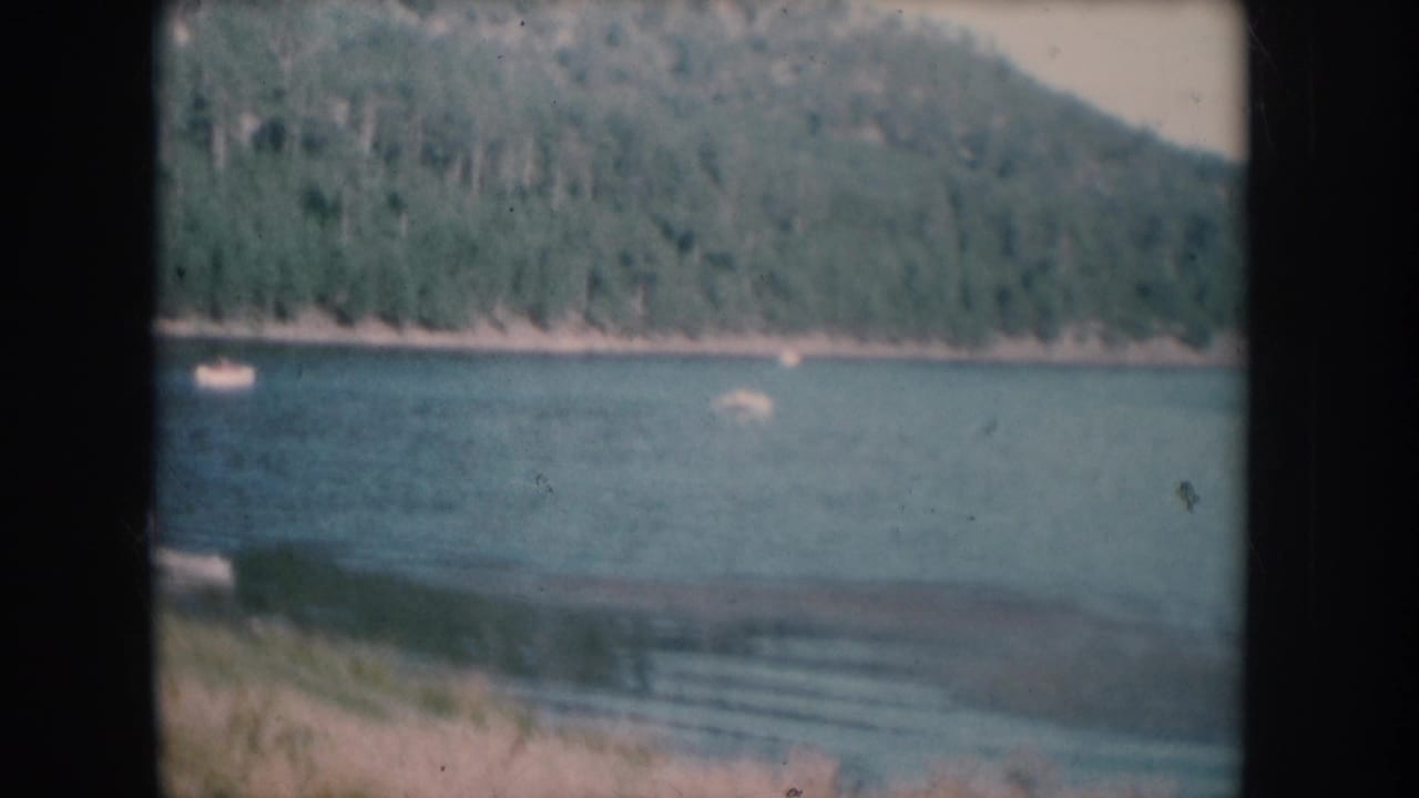 Wide view of a lake with forested hills and a sandy near-shore; small distant figures visible on the water.