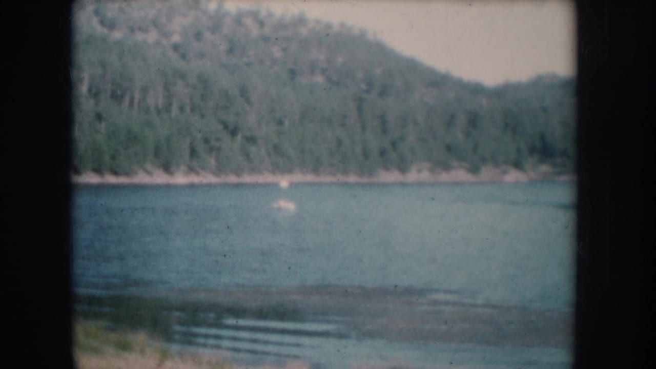 Lake with dense forested hills and a shallow sandy area in the foreground; faint activity visible on the water surface.