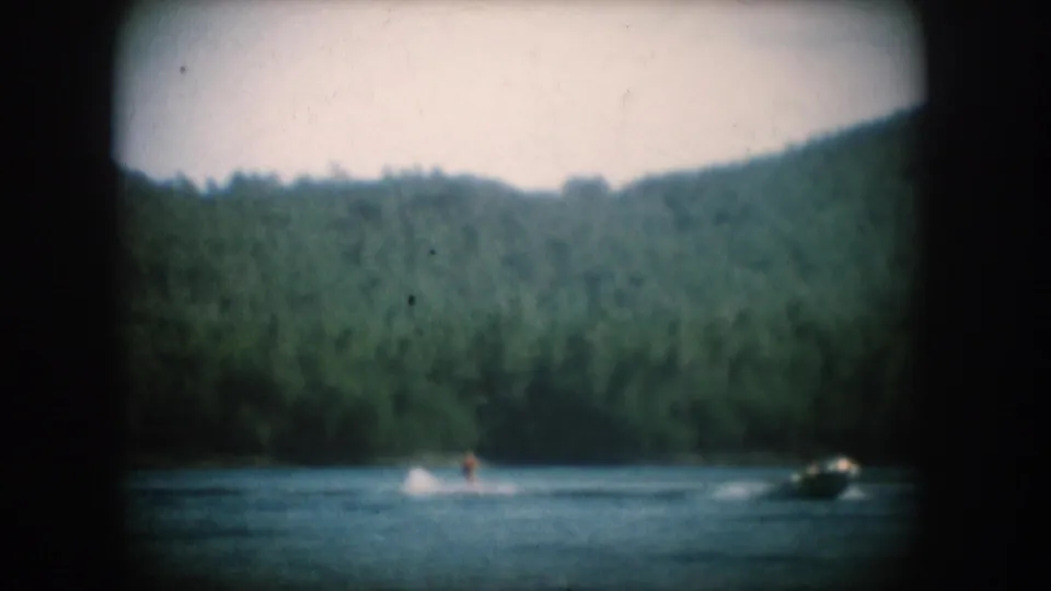 8mm home movie circa 1956, South Dakota lake: a motorboat pulling a water skier below tree-covered hills with a pale overexposed sky above.