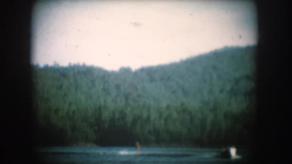 8mm home movie circa 1956, South Dakota: a forested hillside above a calm lake surface with a small distant figure at the water's edge.