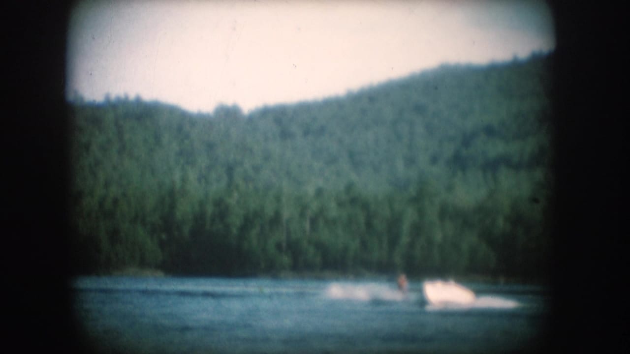Motorboat with white spray wake on a dark lake, forested hill rising across the background, 8mm film.