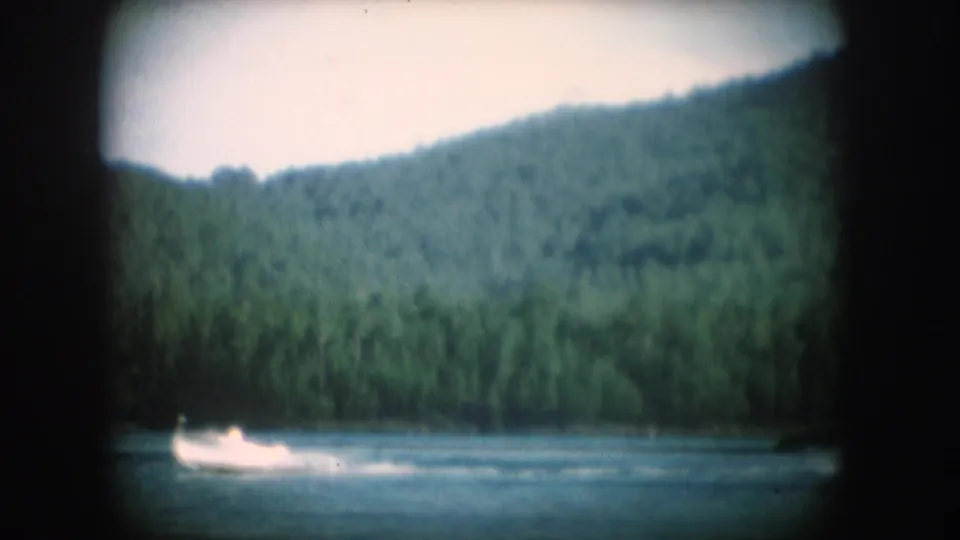 Motorboat trailing a white wake across a forested South Dakota lake, circa 1956, 8mm home movie with pronounced vignetting.
