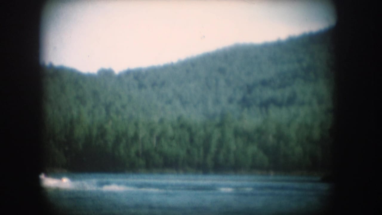1956 8mm home movie — wide lake shot with forested hillside in South Dakota; faint wake or spray visible at lower left.
