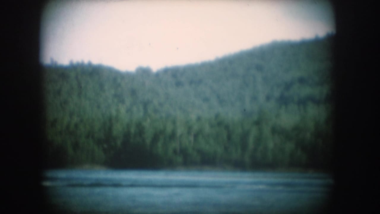 Wide 8mm shot of calm lake and dense forested hillside; water surface undisturbed with no figure visible in frame.