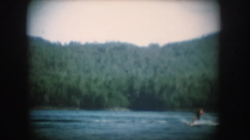 1956 8mm home movie — water skier in dark swimsuit at lower center of South Dakota lake, dense forested hillside behind.