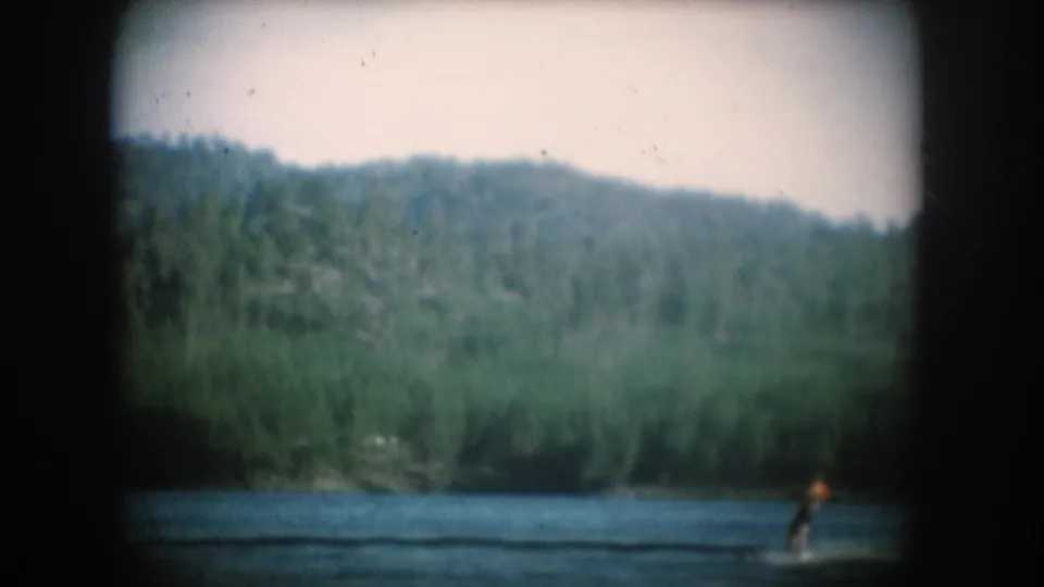 1956 8mm home movie — water skier at lower right of South Dakota lake, dense forested ridge above, film dust specks visible in pale sky.
