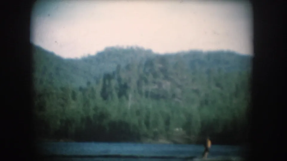 Wide 8mm home-movie frame circa 1956 showing a forested mountainside, calm lake surface, and a small distant figure near the shoreline.