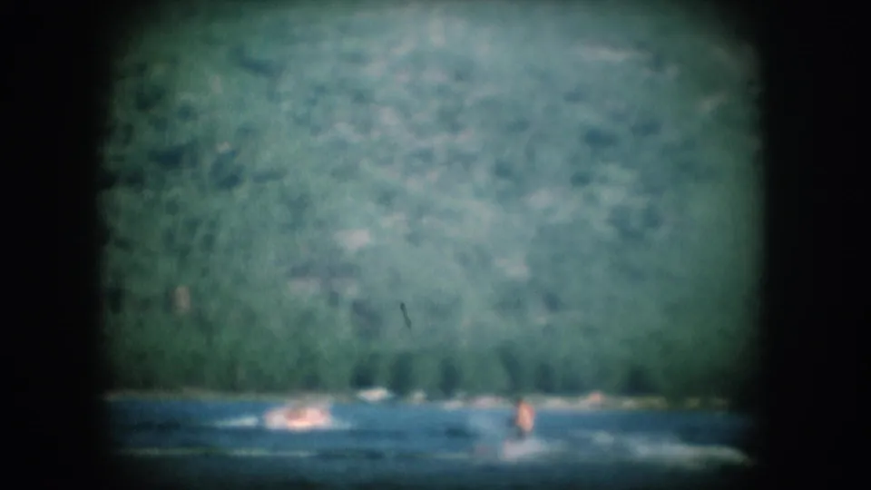 8mm home-movie frame circa 1956 showing water spray and wake with a small figure in motion on a lake, dense forest filling the background.