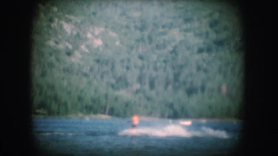 8mm home-movie frame circa 1956 showing a figure amid water spray on a lake, with a densely wooded hillside rising in the background.