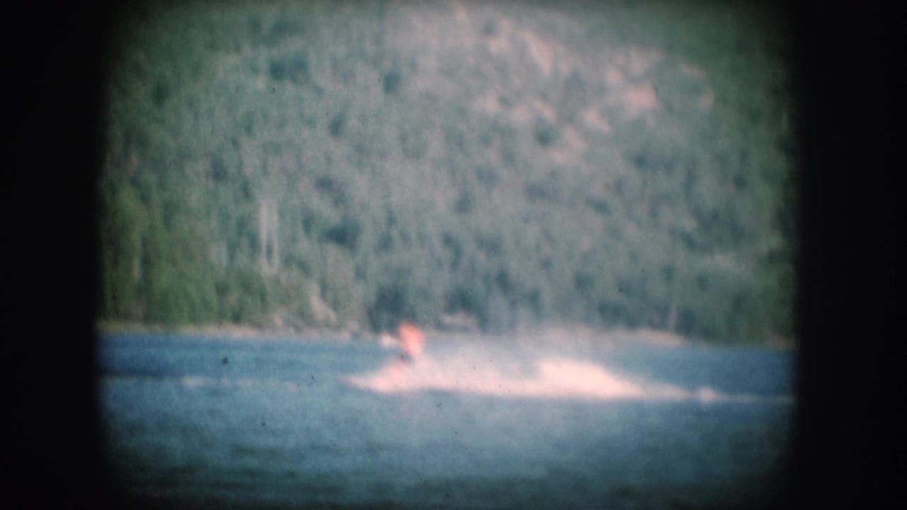 1956 8mm home movie: small figure water skiing on a pine-rimmed lake, likely South Dakota, with faded color palette and film vignetting.