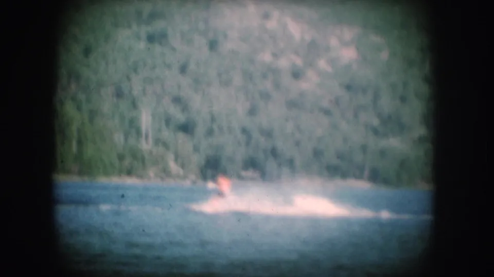 1956 8mm home movie: small figure water skiing on a pine-rimmed lake, likely South Dakota, with faded color palette and film vignetting.