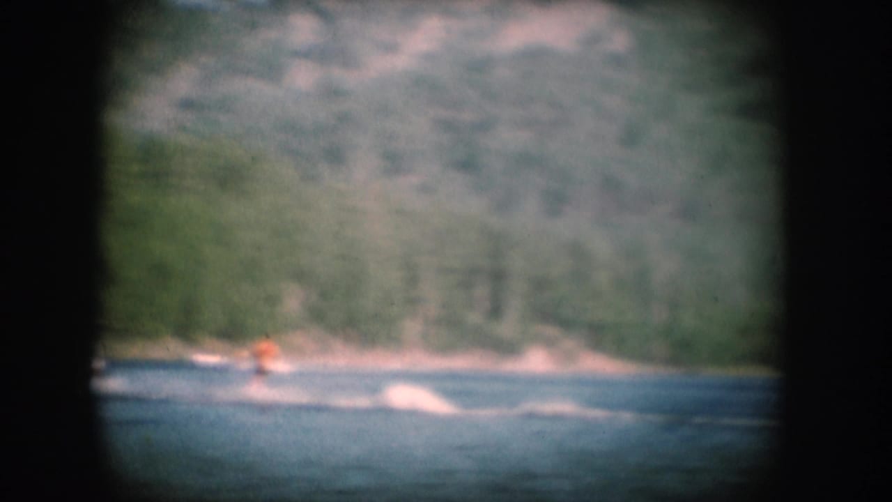 1956 8mm home movie: water skier in orange top crossing a forested lake, likely South Dakota, with film grain and characteristic vignetting.