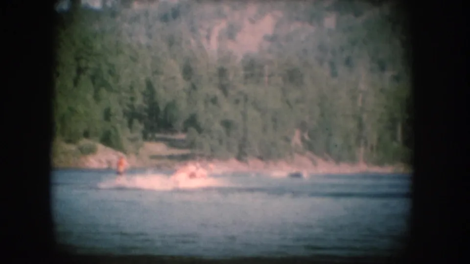 1956 8mm home movie: rocky shoreline with small figures near the water, pine-forested slope rising behind, consistent with a South Dakota lake setting.