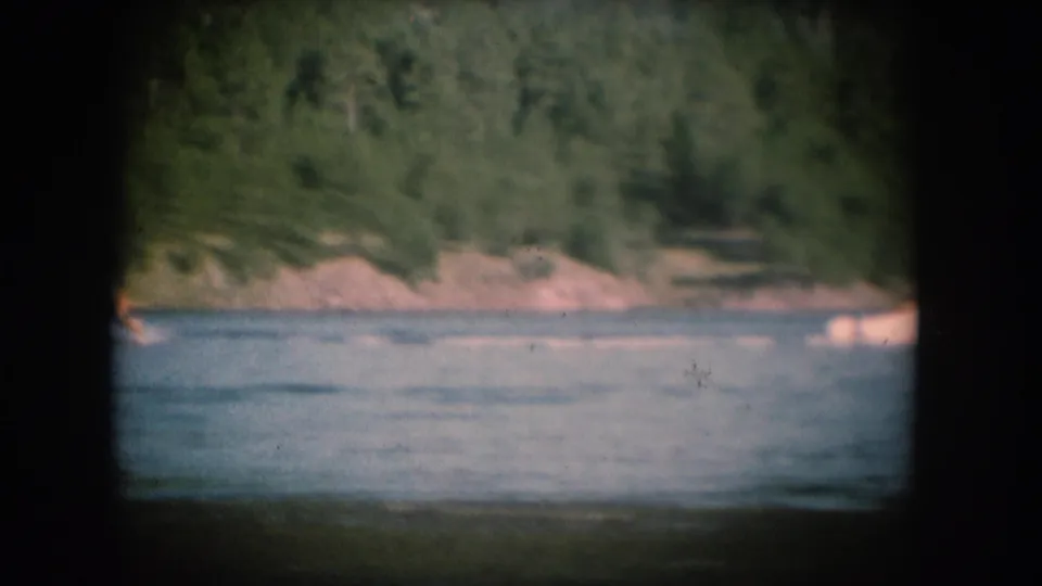 1956 8mm home movie: pine-covered hillside and rocky lakeside with small distant figures, South Dakota setting, with characteristic film grain and vignetting.