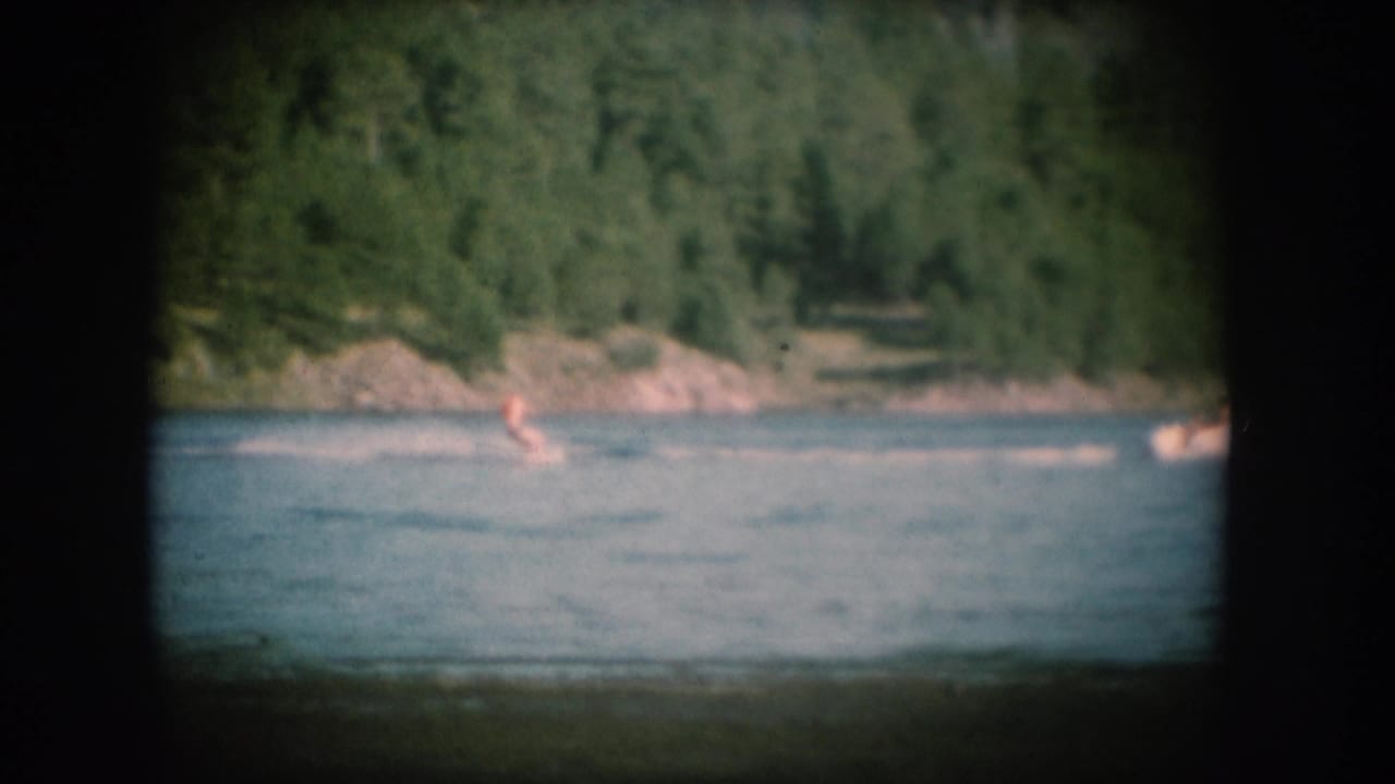 8mm home movie frame, c.1956, showing two water skiers at distance on a blue lake, backed by a steep green forested slope.