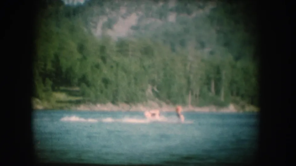 8mm home movie frame, c.1956, showing a lone figure at distance on a blue lake with a dense forest-covered slope behind.
