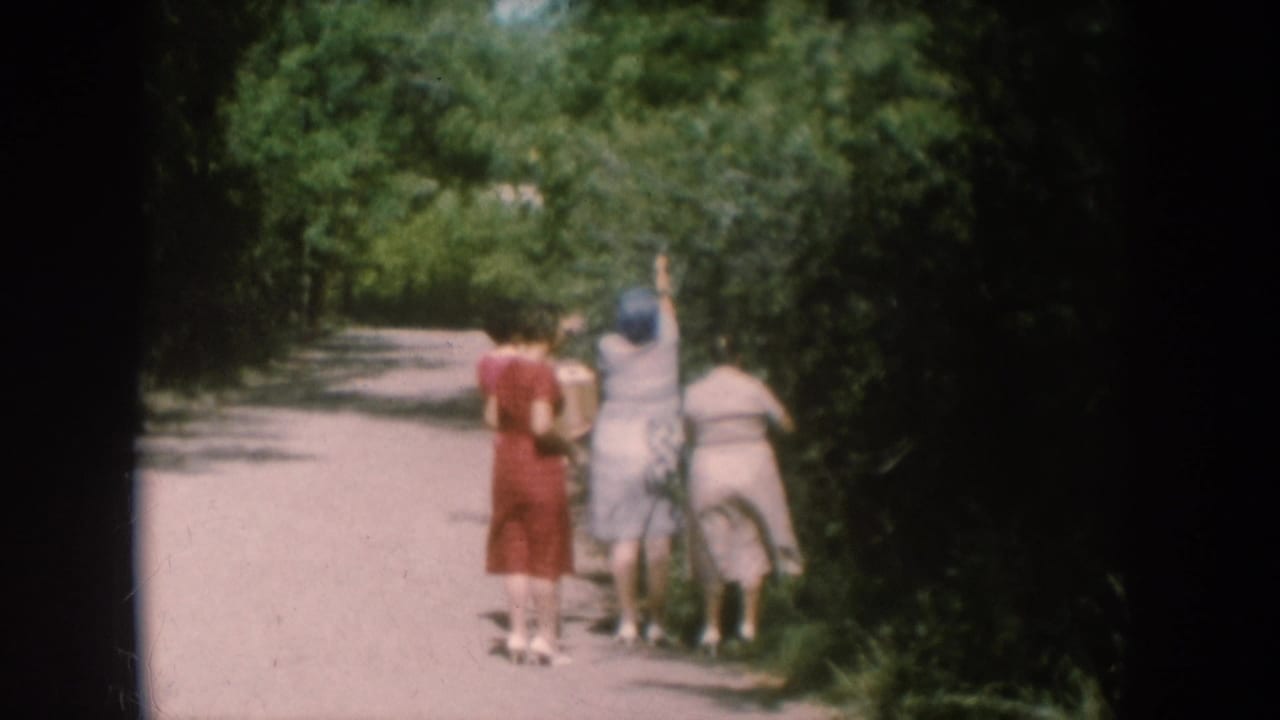 Three women approaching a berry bush along a rural dirt path, South Dakota, circa 1956; red and light-colored summer dresses, lush green foliage.