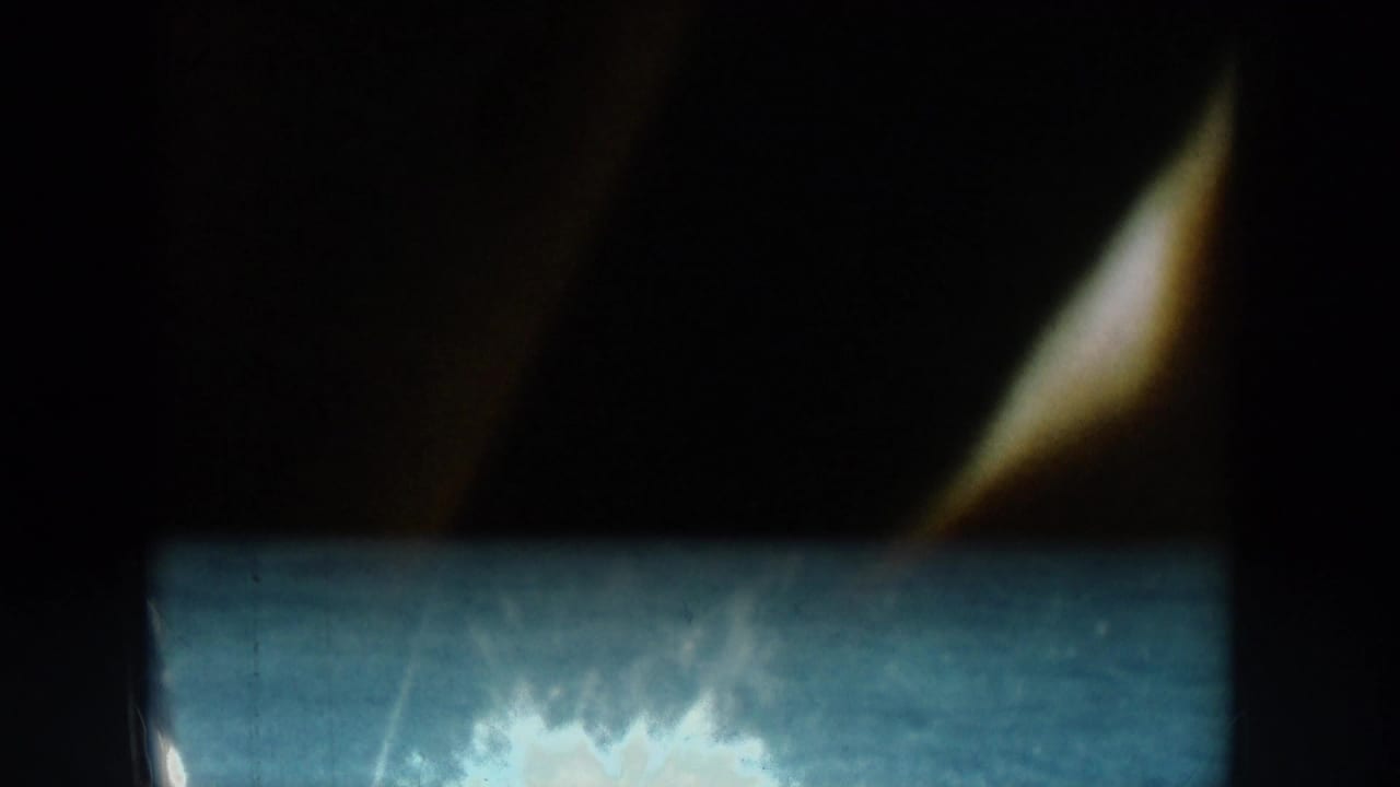 Opening frame of an 8mm home movie reel, largely black with a brief burst of reflected light or spray suggesting the camera was still panning or starting.