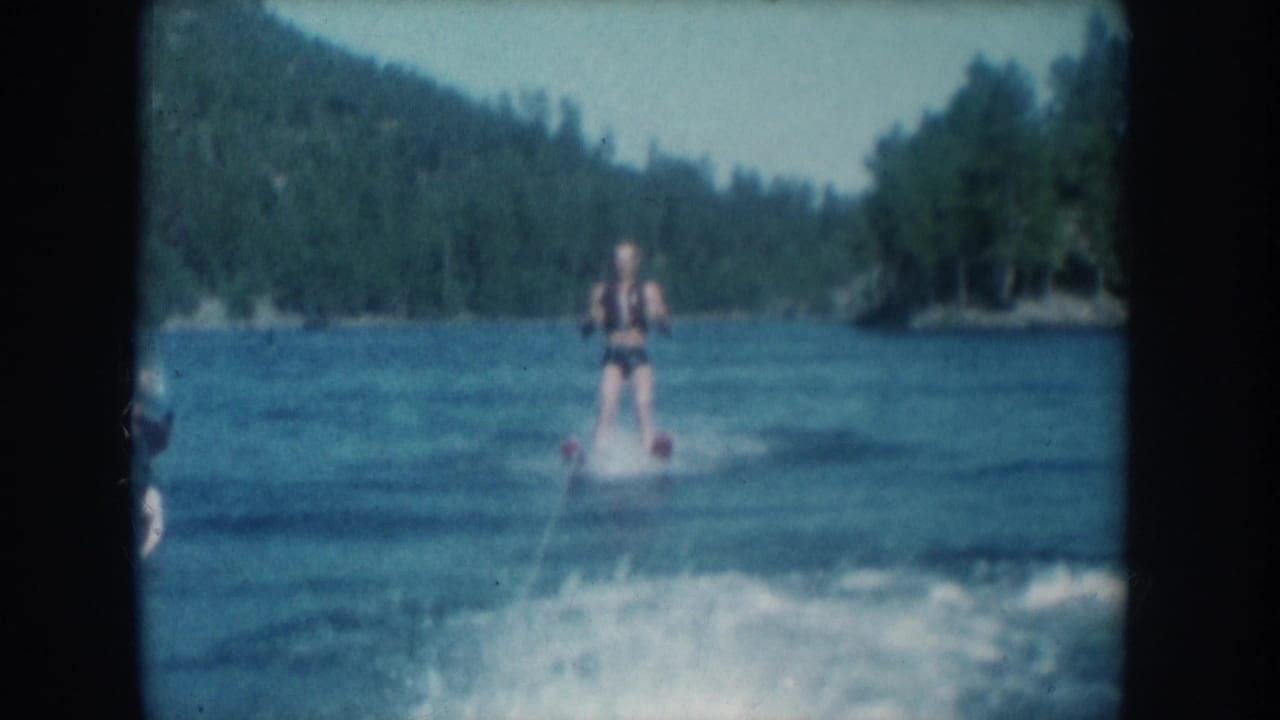 Water skier in life vest holds a tow rope upright on a blue lake; dense coniferous hillside fills the background.