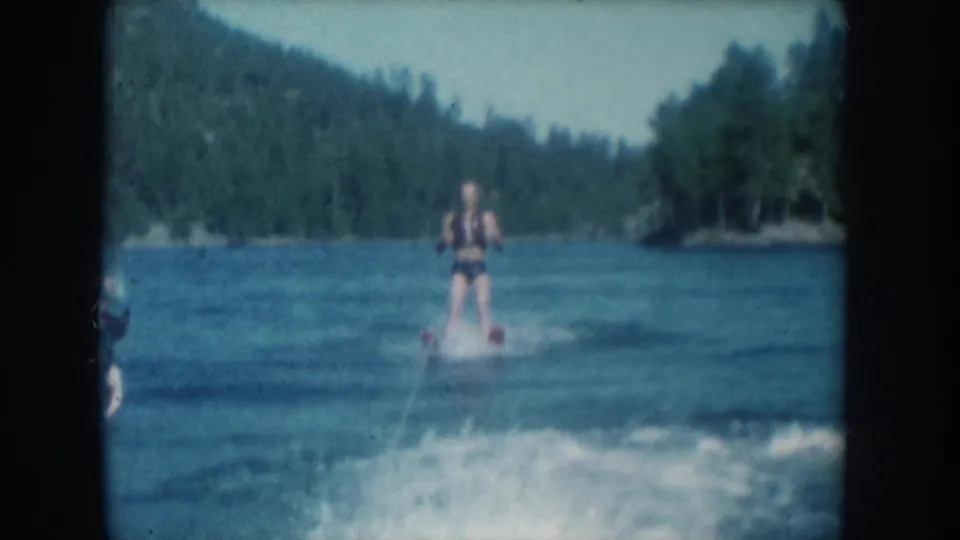 Mid-1950s 8mm home movie frame of a water skier on a forested lake, metadata-attributed to South Dakota, USA, 1956; skier wears a life vest and holds a tow rope.