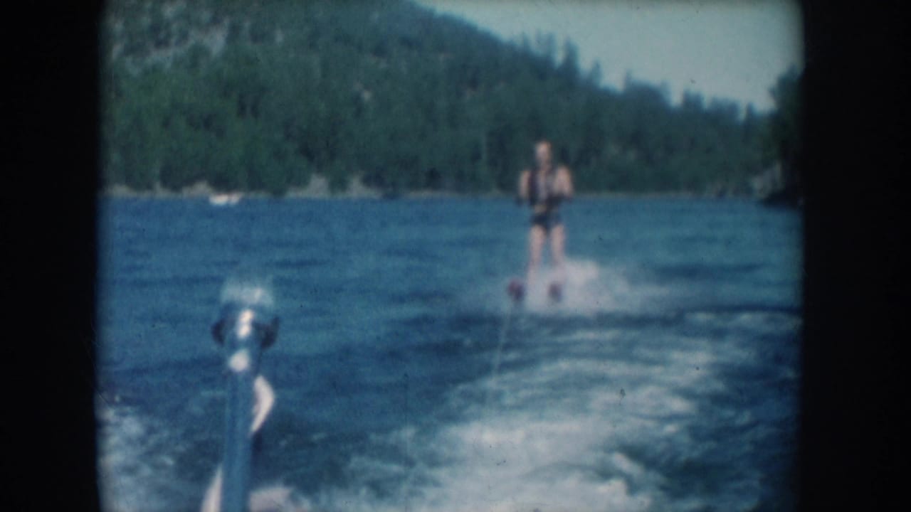 8mm home movie frame shot from aboard the towing boat looking back at a water skier on a forested lake, circa 1956, South Dakota; outboard motor partially visible at lower left.