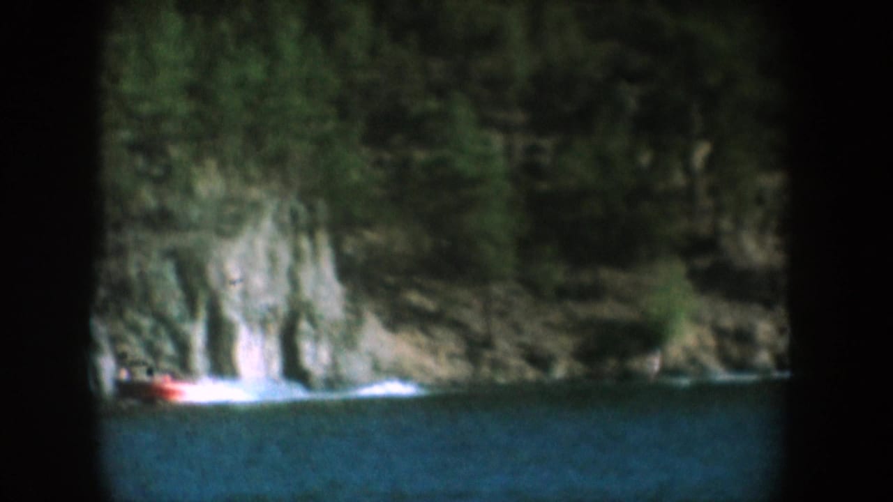 Rugged stone cliff descending to still lake water with a small red boat visible at the base; pine trees crown the cliff; filmed on 8mm circa 1956 in what metadata identifies as South Dakota.