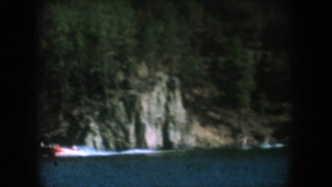 Red motorboat crossing a lake backed by a grey rock face and pine forest, attributed to South Dakota, circa 1956, 8mm home movie.