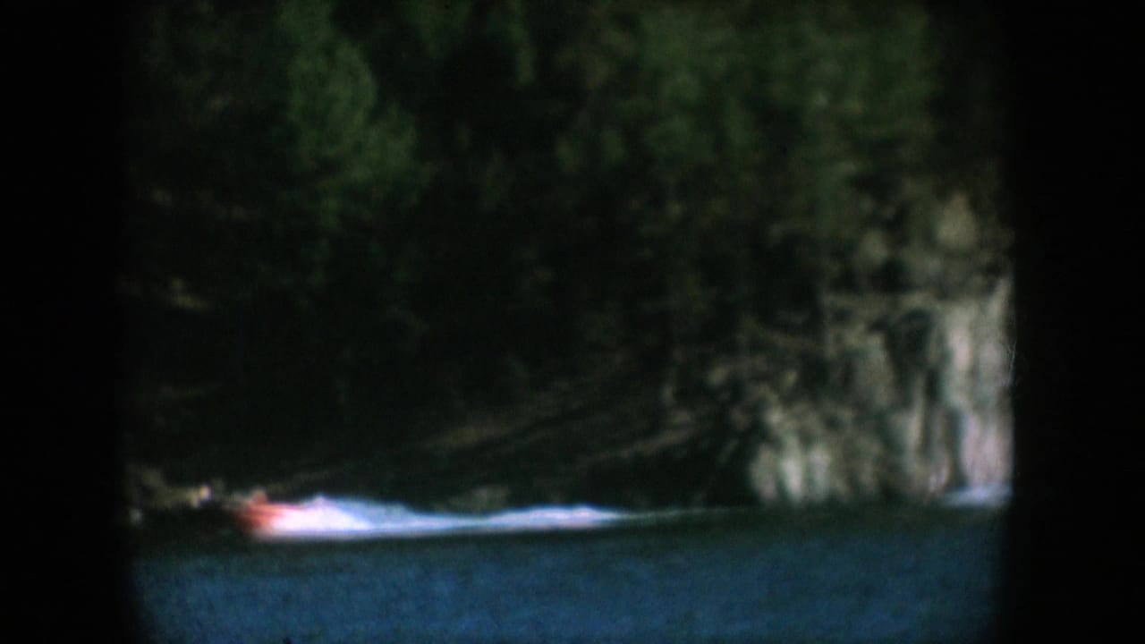 Red motorboat travelling along a forest-lined lake with a rock face at centre, South Dakota, circa 1956, 8mm home movie.
