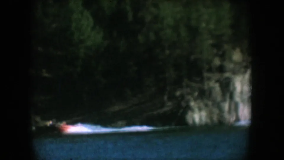 Red motorboat travelling along a forest-lined lake with a rock face at centre, South Dakota, circa 1956, 8mm home movie.