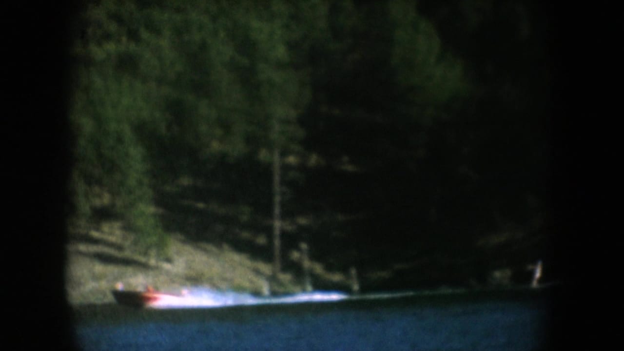 Red motorboat passing a forested lakeshore with sandy bank visible after camera pans from cliff face, South Dakota, circa 1956, 8mm.