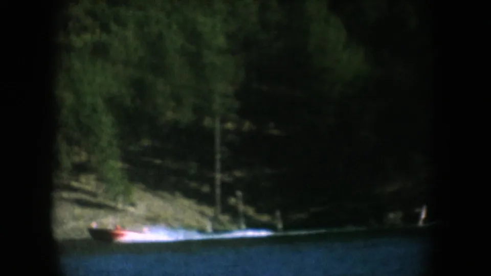 Red motorboat passing a forested lakeshore with sandy bank visible after camera pans from cliff face, South Dakota, circa 1956, 8mm.