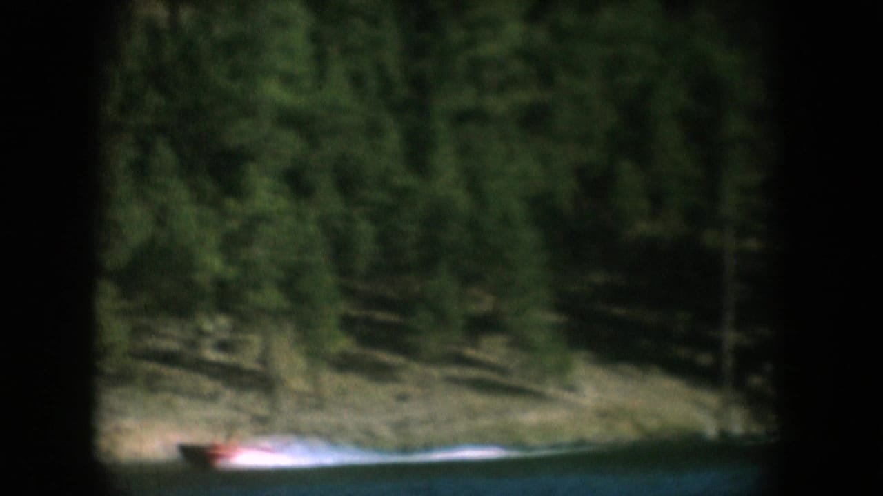 Red motorboat receding toward the far shore of a forested lake, sandy bank and pine trees visible, South Dakota, circa 1956, 8mm home movie.