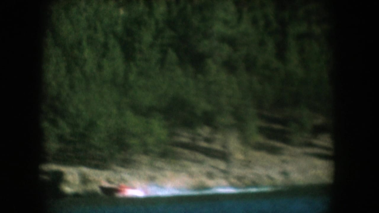 Small red speedboat crossing a forested lake, captured on 8mm film, likely South Dakota, circa 1956.