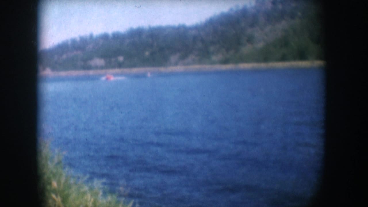 8mm home movie frame circa 1956, South Dakota lake — distant water skier in red top, tow boat, conifer-covered hillside, characteristic gate vignette border.