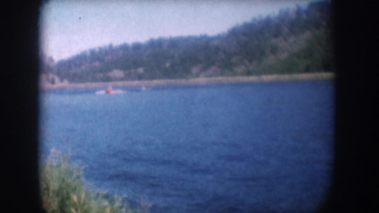 Blue lake with distant water skier in red top and tow boat; dark conifer hillside along far shore; 8mm dark border visible.