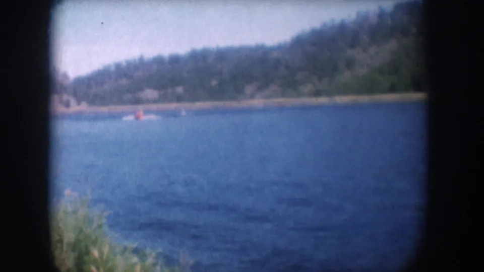 8mm home movie frame circa 1956, South Dakota lake — skier with red top still visible mid-water, dense forested hillside, pale summer sky above.