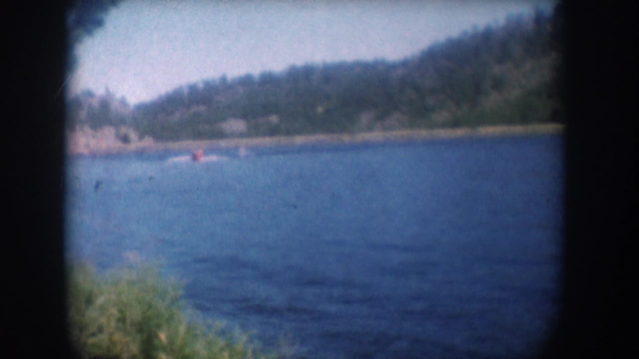 8mm home movie frame circa 1956, South Dakota lake — water skier near rocky far shoreline, pine-covered hillside, what may be a lakeshore structure in the far distance.