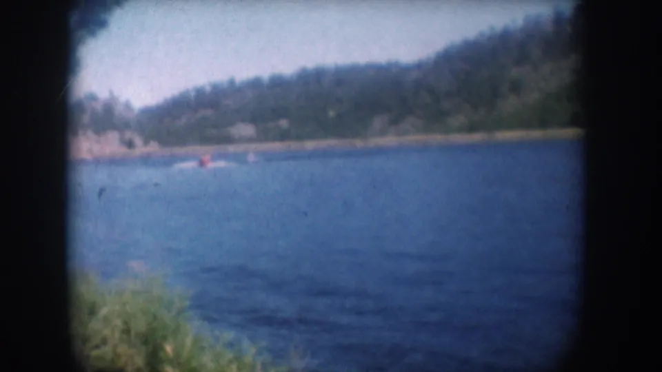 8mm home movie frame circa 1956, South Dakota lake — water skier near rocky far shoreline, pine-covered hillside, what may be a lakeshore structure in the far distance.