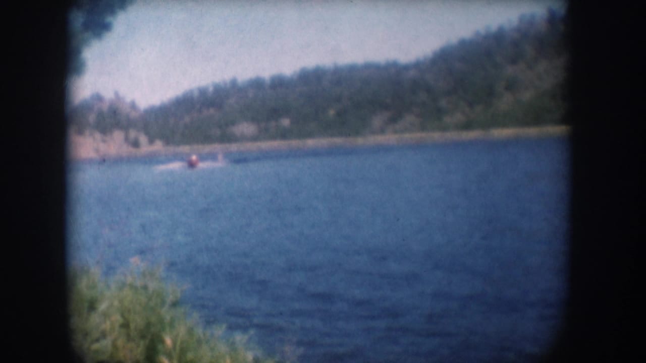 8mm home movie frame circa 1956, South Dakota lake — water skier continuing across lake, pine hillside and rocky shore in background, warm faded mid-century color.