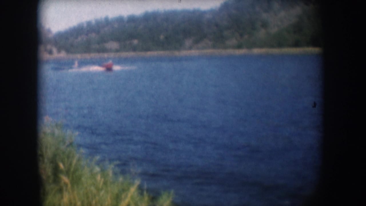 Red motorboat towing a water skier across a South Dakota lake circa 1956, with green shore reeds at left and a wooded hillside beyond, in 8mm home movie footage.