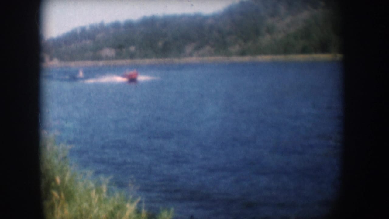 Red motorboat crossing a South Dakota lake circa 1956, with a faint trailing skier, wooded far shore, and pale sky; 8mm home movie footage with visible film grain.