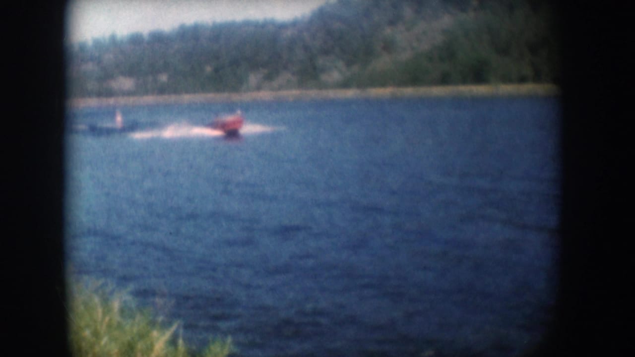 Red motorboat with prominent wake and trailing skier on a South Dakota lake circa 1956; green reeds at lower left and wooded hillside behind, in 8mm home movie footage.