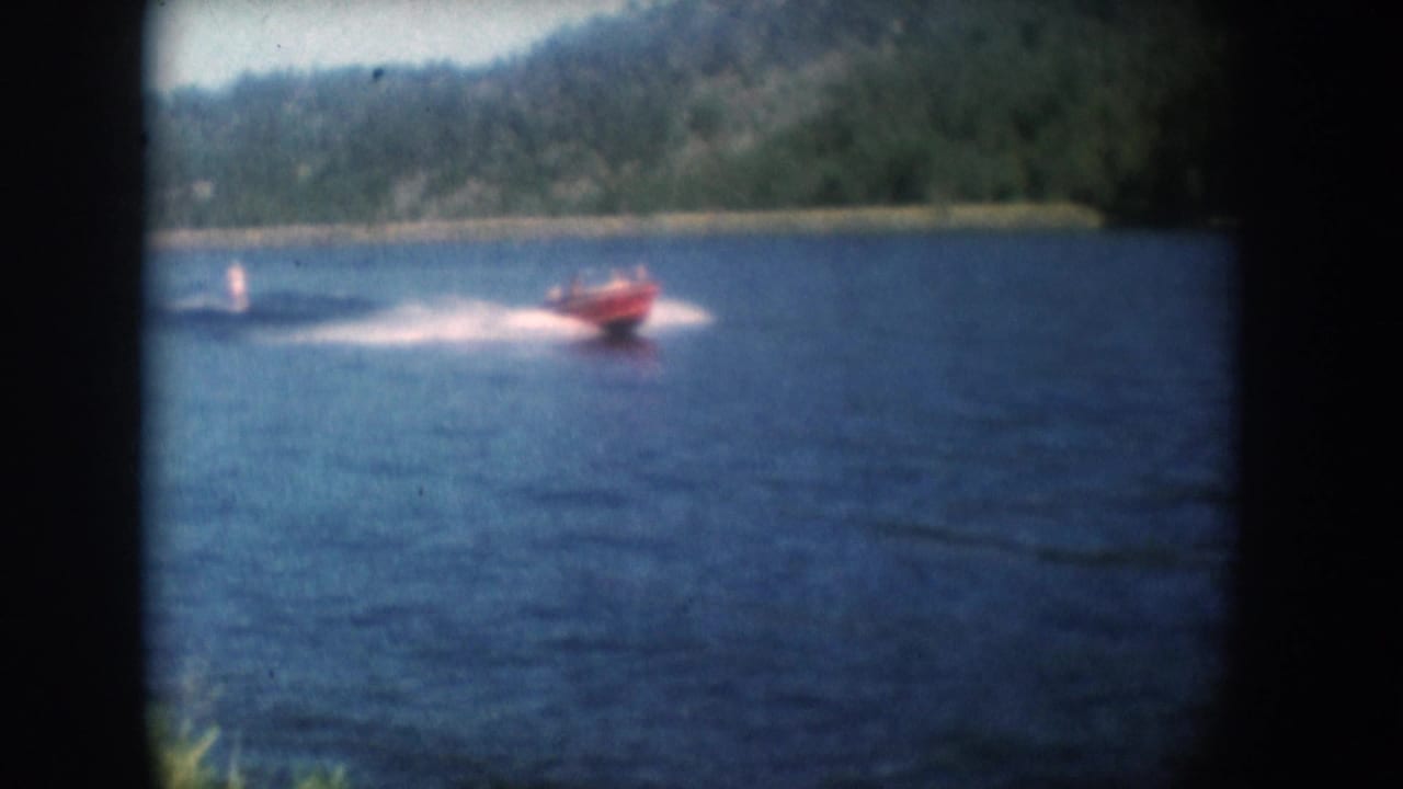 Red motorboat and distant water skier on a South Dakota lake circa 1956; wide wake spray prominent in this stationary-camera 8mm home movie shot with wooded hills beyond.
