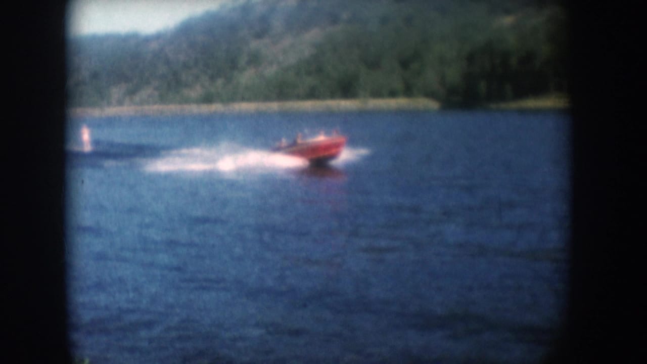 Red motorboat pulling a water skier across a South Dakota lake in 1956, with a wooded hillside beyond, shot on 8mm color film.